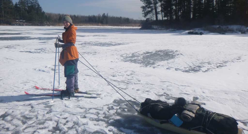 A person stands on cross country skis with a small sled attached by poles at their waste. They appear to be standing on a frozen lake. 